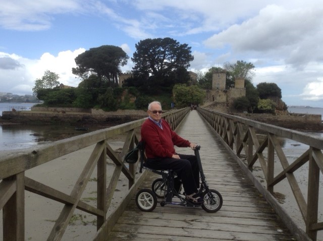 A man sat on a Di Blasi (Stowaway Morphic) mobility scooter on a walkway towards an island with his